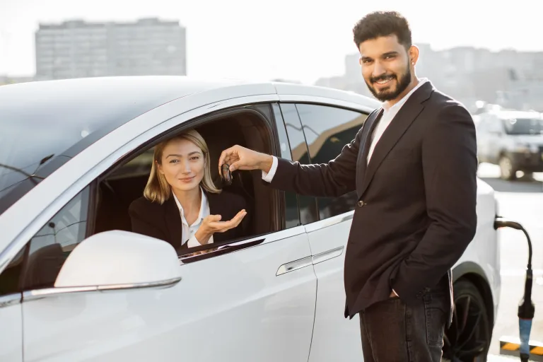 A smiling car rental agent hands over the keys to a businesswoman sitting inside a white luxury car. The vehicle is parked outdoors with a modern cityscape in the background, representing premium rent-a-car services in Abu Dhabi and Dubai.